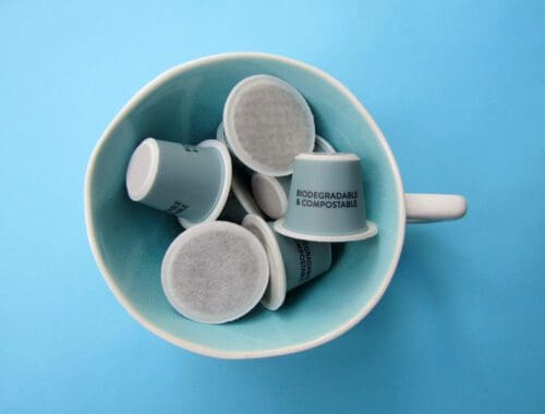 A white cup filled with several light blue coffee pods, labeled "Biodegradable & Compostable," is placed against a matching light blue background. These innovative coffee pods aim to eliminate waste and promote sustainability.