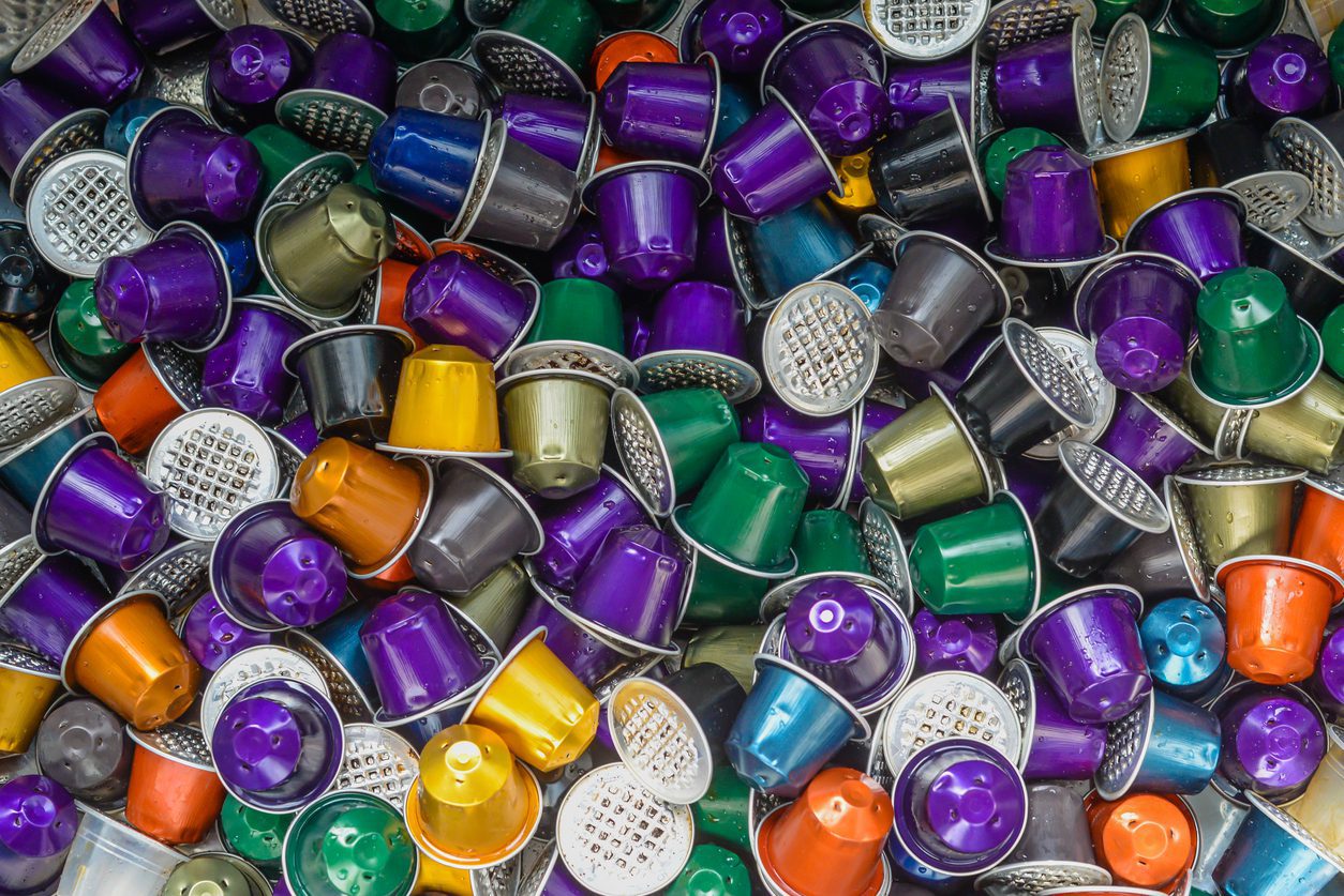 A large collection of colorful coffee pods in purple, yellow, green, orange, blue, and silver are piled together. The pods have a shiny metallic appearance and are designed for use in single-serve coffee machines.