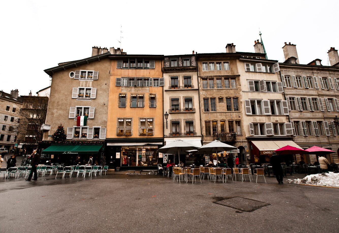 A quaint street scene showcasing several historic buildings with varied facades and shuttered windows. Outdoor cafes with tables, chairs, and colorful umbrellas line the sidewalk. People are casually walking and seated in the area, savoring their drinks made with organic coffee beans, enjoying the atmosphere.