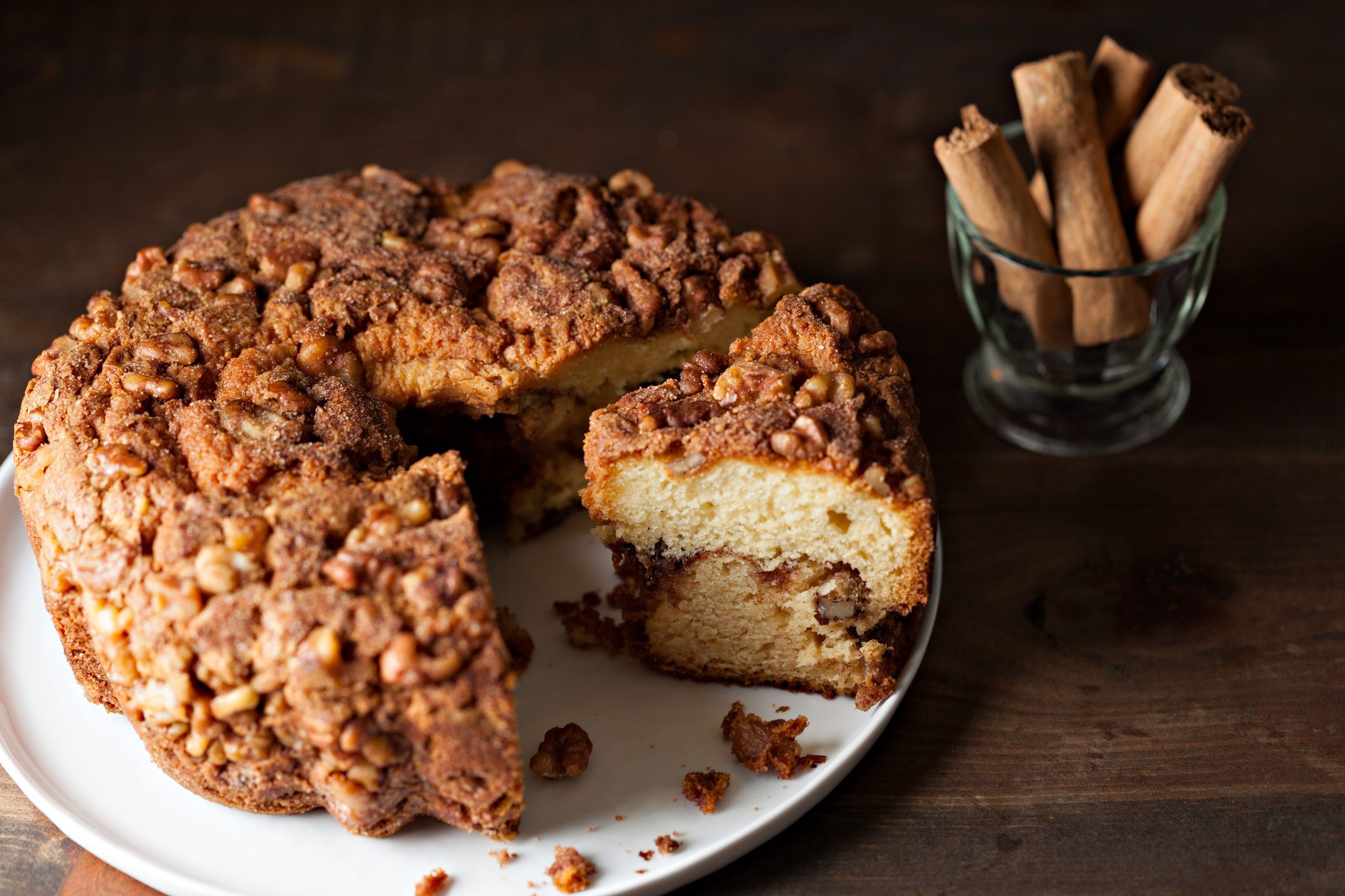 A round cinnamon coffee cake with a slice removed, revealing a marbled interior. The cake is topped with a crumbly streusel and chopped nuts, providing not just flavor but functional benefits. Next to it, a small, clear glass holds several cinnamon sticks. Everything is set against a dark wooden background.