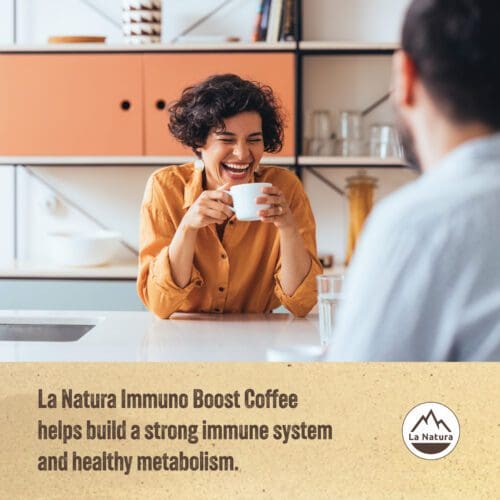 A person with short, curly hair and wearing a mustard-colored shirt sits at a kitchen counter, smiling and holding a cup of coffee. The text on the image says, "La Natura Immuno Boost Coffee Pods - Crema Roast helps build a strong immune system and healthy metabolism.