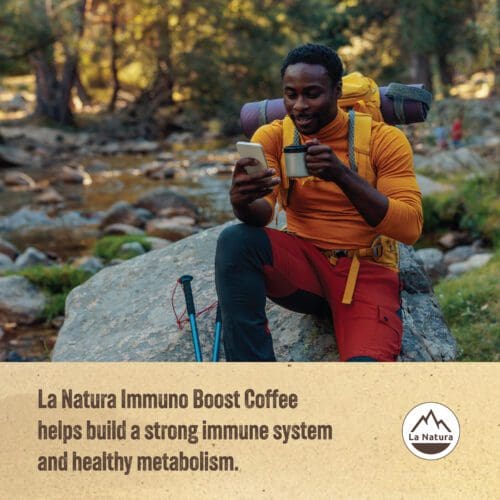 A hiker sits on a rock near a stream, dressed in outdoor gear, with a backpack beside him. He is smiling at his phone while holding a mug of coffee. The text reads, "La Natura Immuno Boost Coffee Pods - Crema Roast helps build a strong immune system and healthy metabolism with every creamy sip.