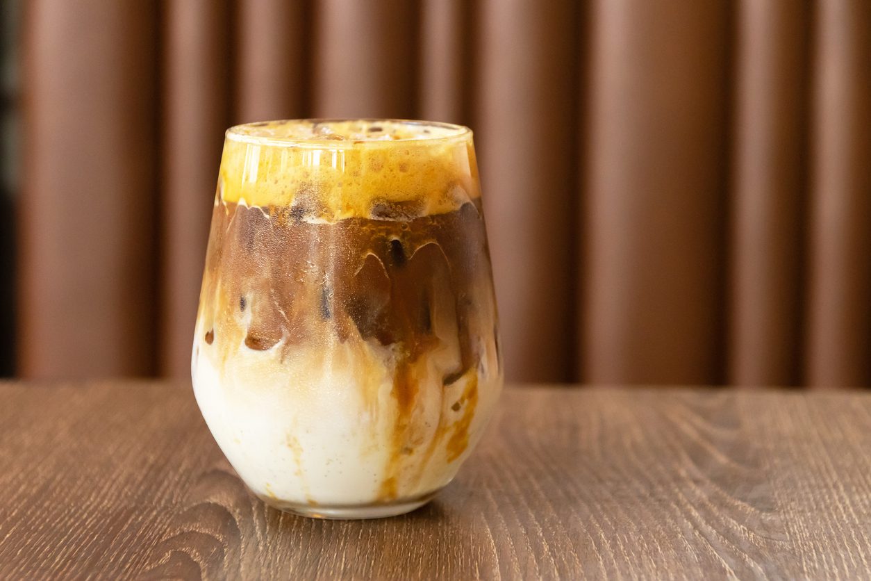 A close-up image of a glass filled with a layered iced coffee drink. The drink features milk at the bottom, a middle layer of coffee, and foam on top. Swirls of coffee and milk blend together along the sides of the glass, reminiscent of popular summer coffee recipes. The background is blurred brown.