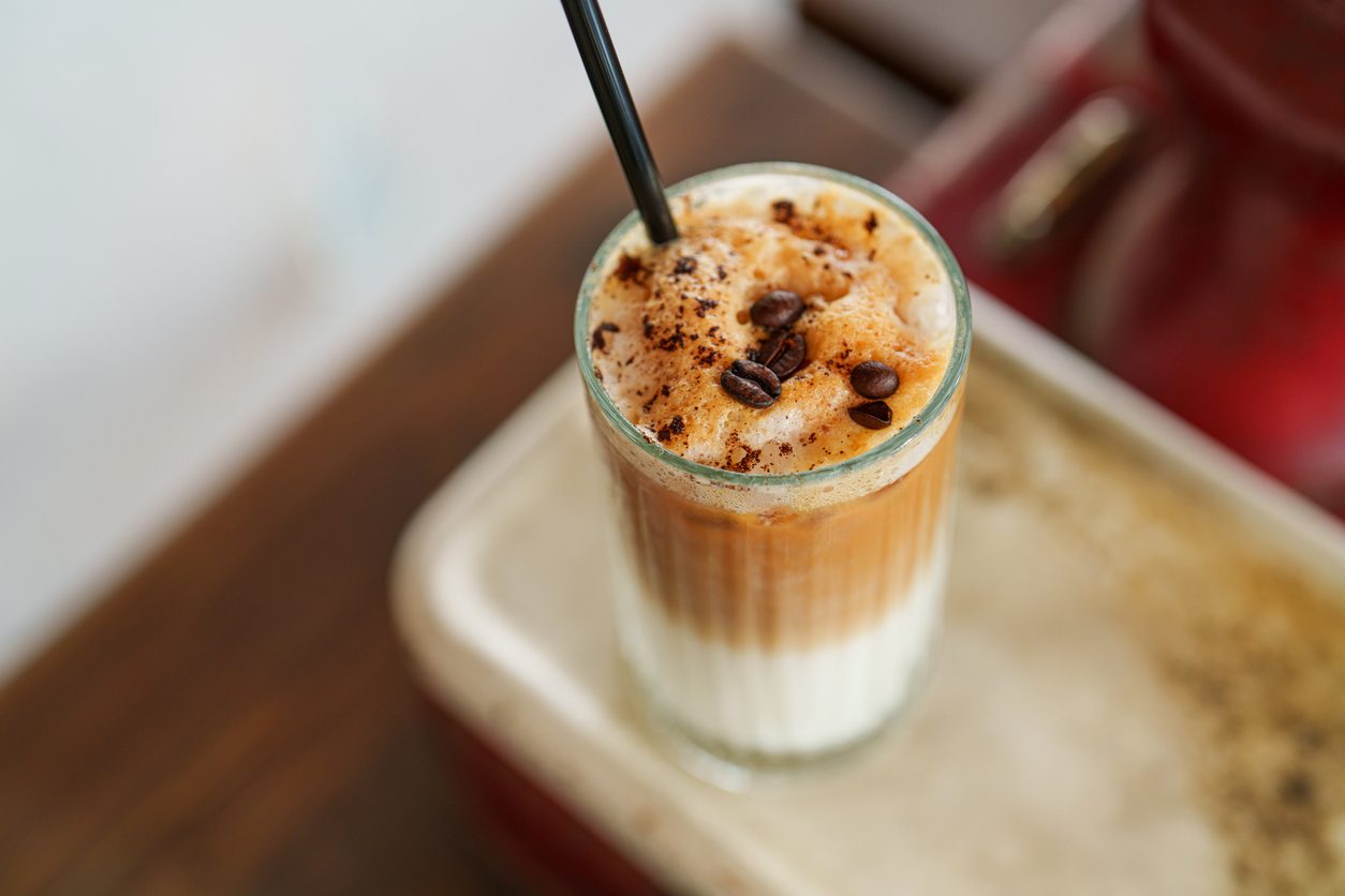 A glass of iced coffee topped with frothy milk, coffee beans, and a sprinkling of cocoa powder. A black straw is inserted into the drink. The glass is placed on a tray on a wooden surface, perfect for summer coffee recipes from La Natura Coffee. The background includes a blurred red object.