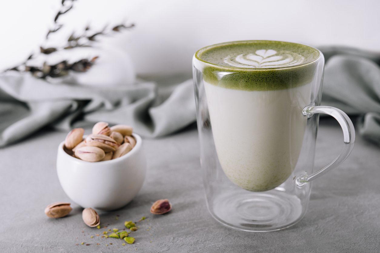 A clear glass mug of matcha latte with a latte art heart on top rests on a gray surface. Beside it, a small white bowl filled with pistachios adds charm, while gray fabric and minimalist decor enhance the calm ambiance. Enjoy this serene setup with La Natura Coffee's healthy coffee blends for a perfect touch.