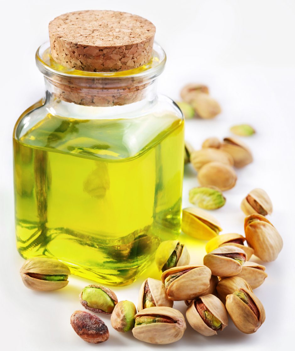 A small glass bottle with a cork stopper contains light green pistachio oil. Surrounding the bottle are scattered pistachios, some shelled and some unshelled, on a white surface—perfect for creating an Iced Pistachio Latte or adding to your homemade coffee syrup.